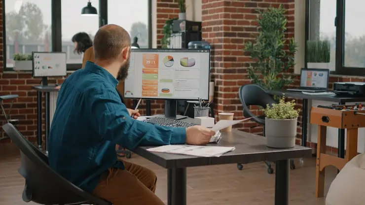 A man working at a desk with charts on his monitor in a brick-walled office